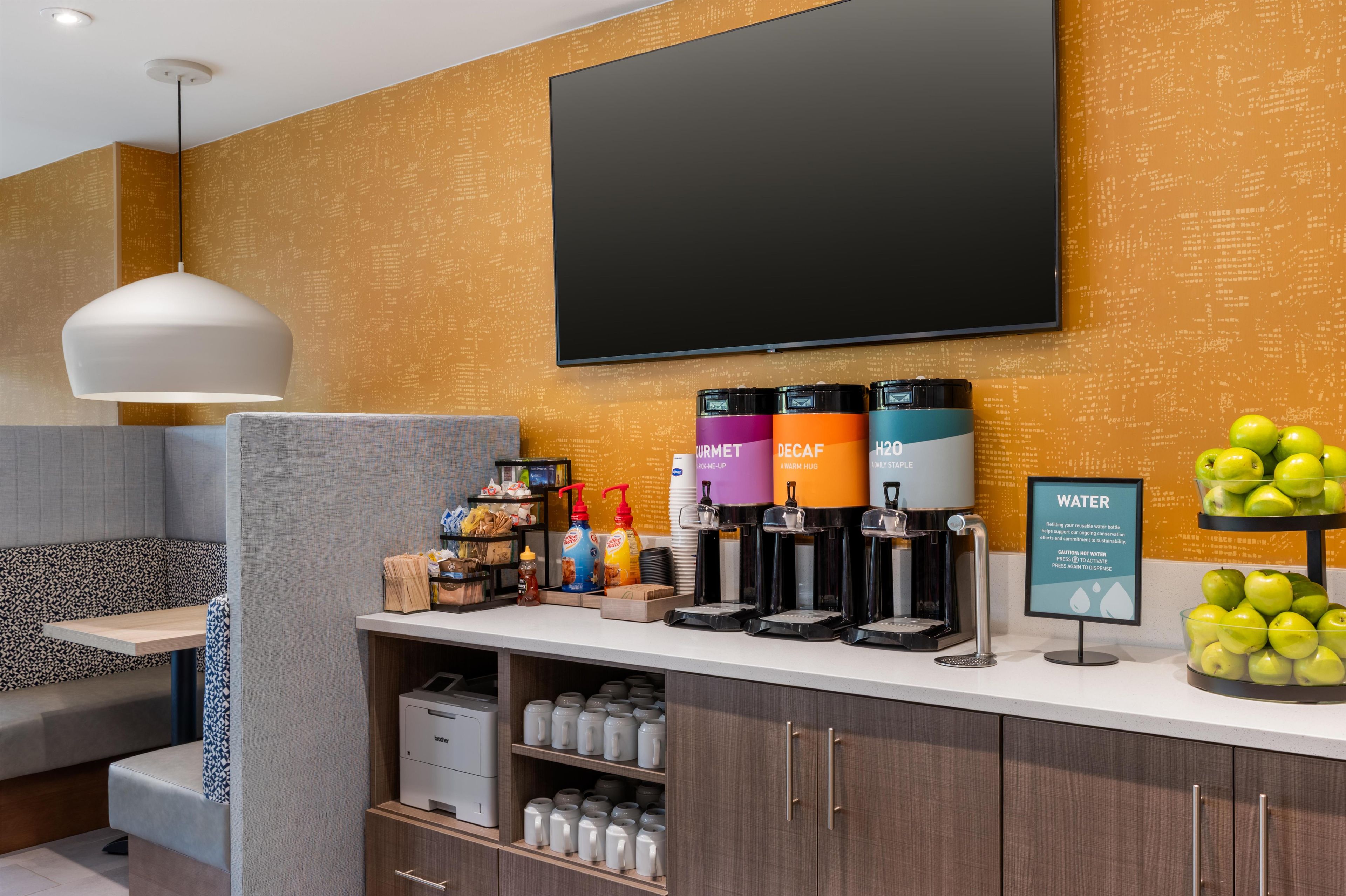 A hotel reception area with a flat-screen TV, coffee machines, a water dispenser, and a fruit display.