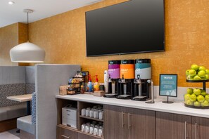 Ein Hotel-Rezeptionsbereich mit Flachbildfernseher, Kaffeeautomaten, Wasserspender und einer Obstauslage.
