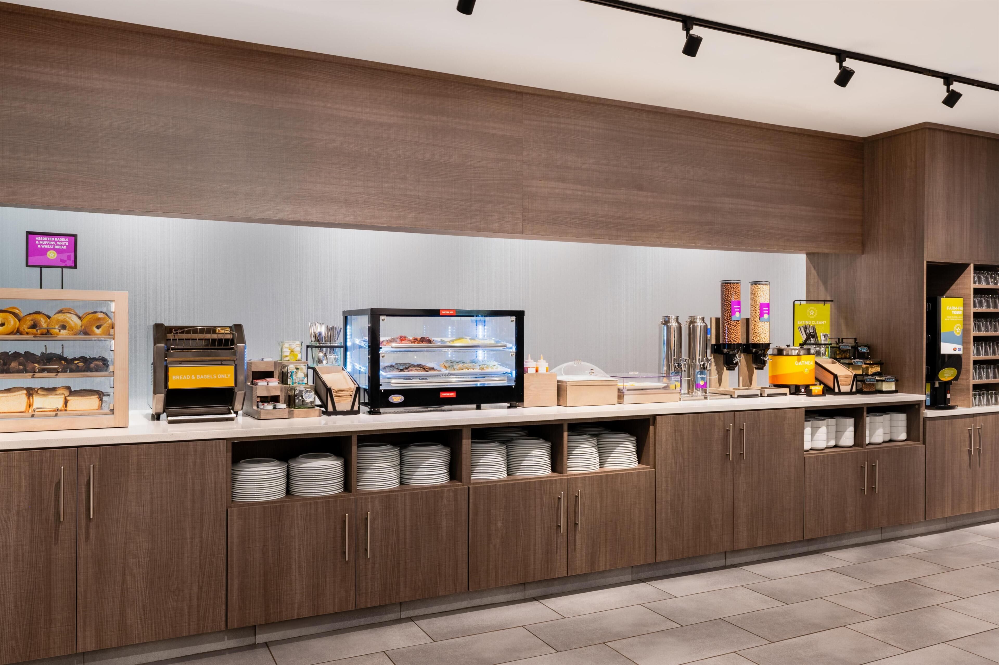 A self-service breakfast station with a digital menu display, bread and bagels, and a coffee machine.