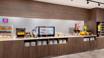 Un mostrador de desayuno autoservicio con menú digital, pan y bagels, y una cafetera.