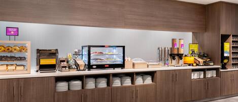 A self-service breakfast station with a digital menu display, bread and bagels, and a coffee machine.