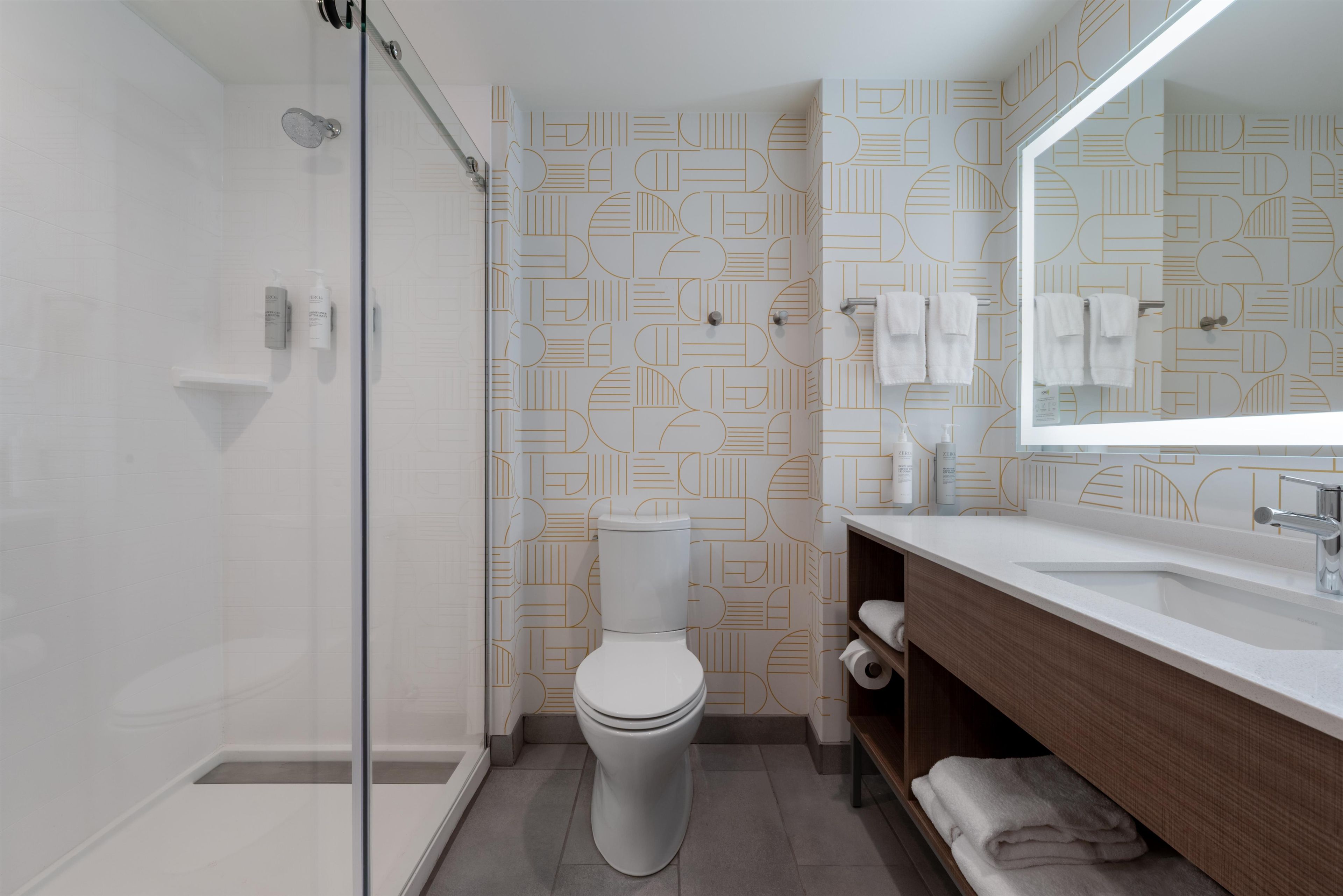 A modern bathroom with a glass shower enclosure, a toilet, a sink with a mirror, and patterned wall tiles.