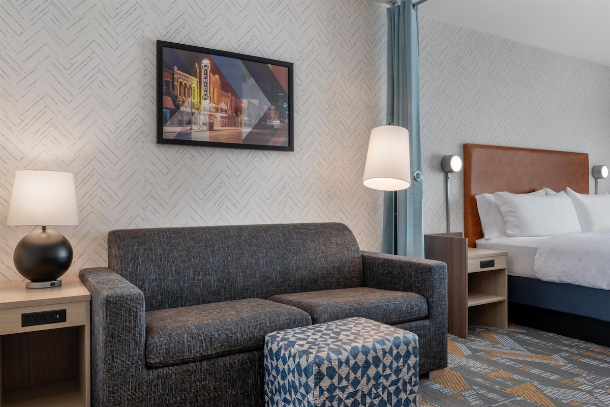 Una habitación de hotel con sofá, reposapiés, mesita de noche, lámpara, mesita auxiliar y una cama con cabecero.
