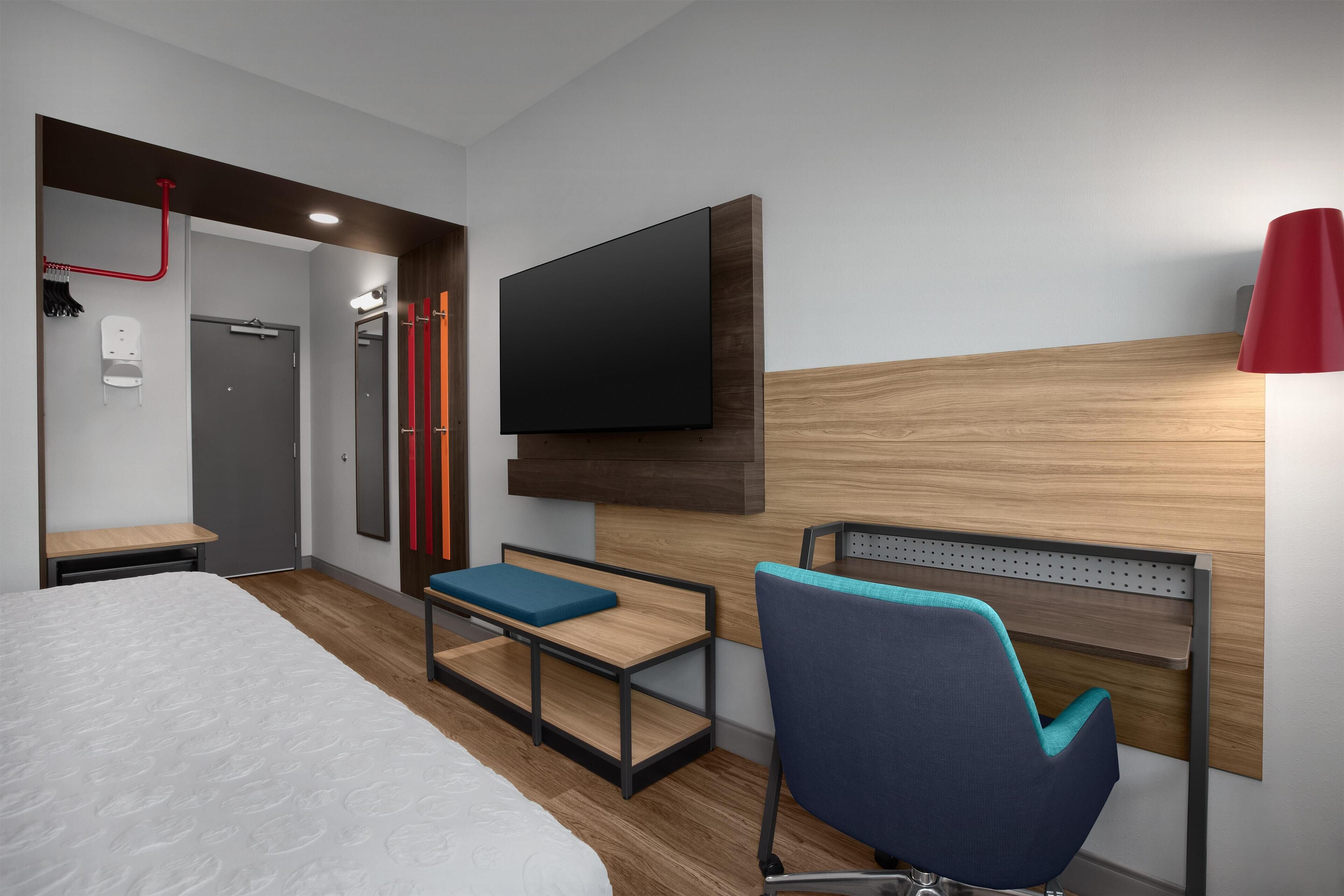 A modern hotel room with a bed, a desk, a chair, a TV, and a red lamp.