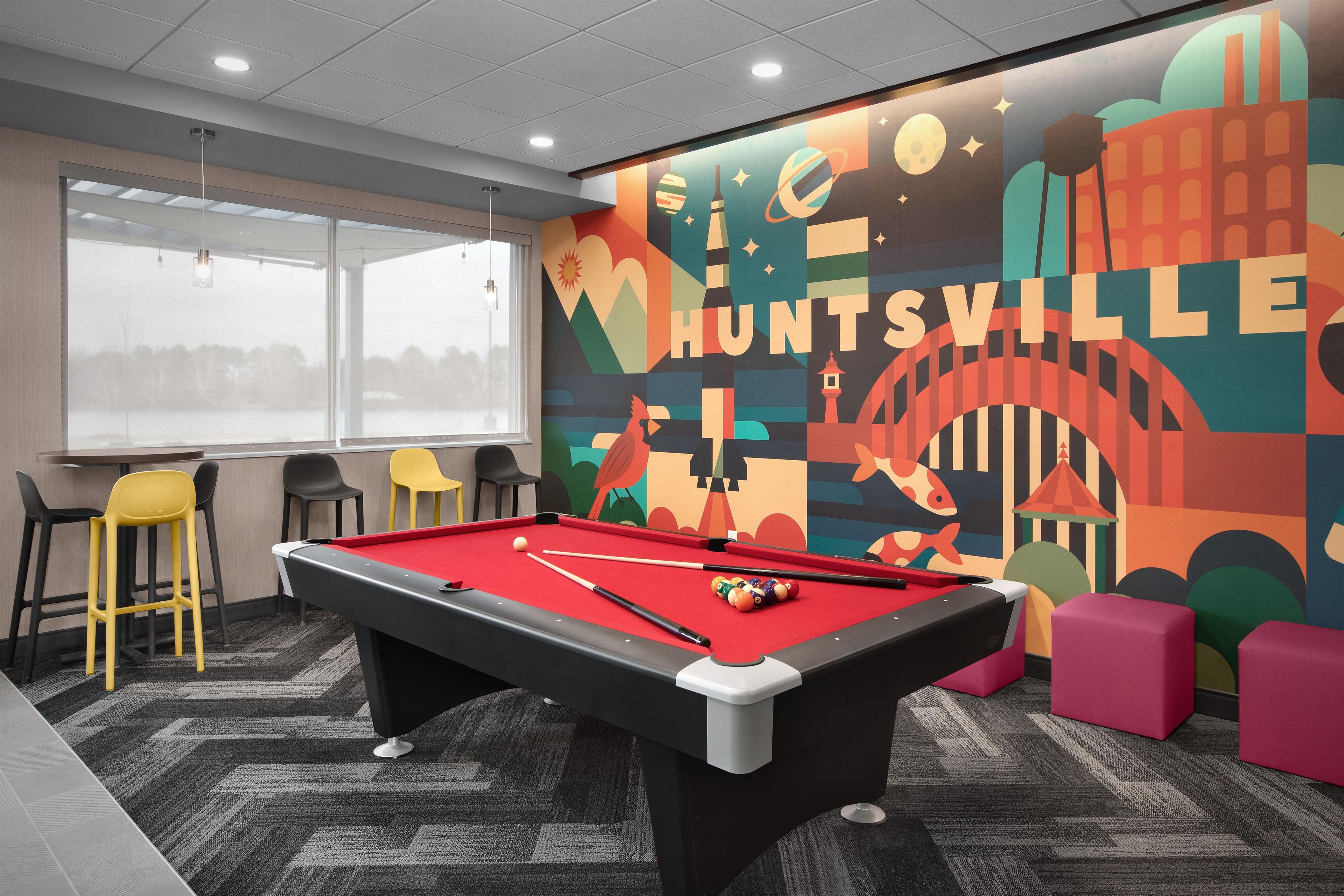 Une table de billard dans une pièce décorée d’une fresque murale colorée, avec des chaises et une fenêtre.