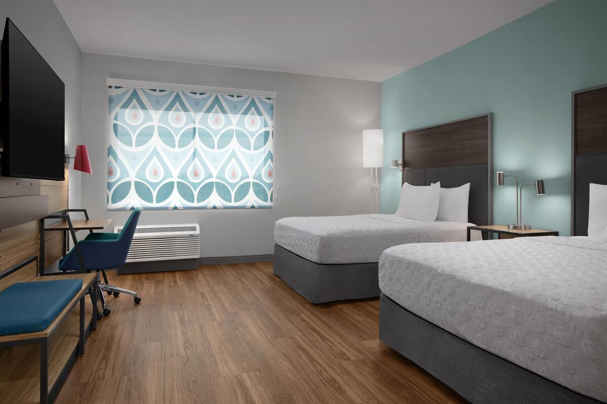 A hotel room with two beds, a desk, a chair, a TV, and a window with a patterned curtain.