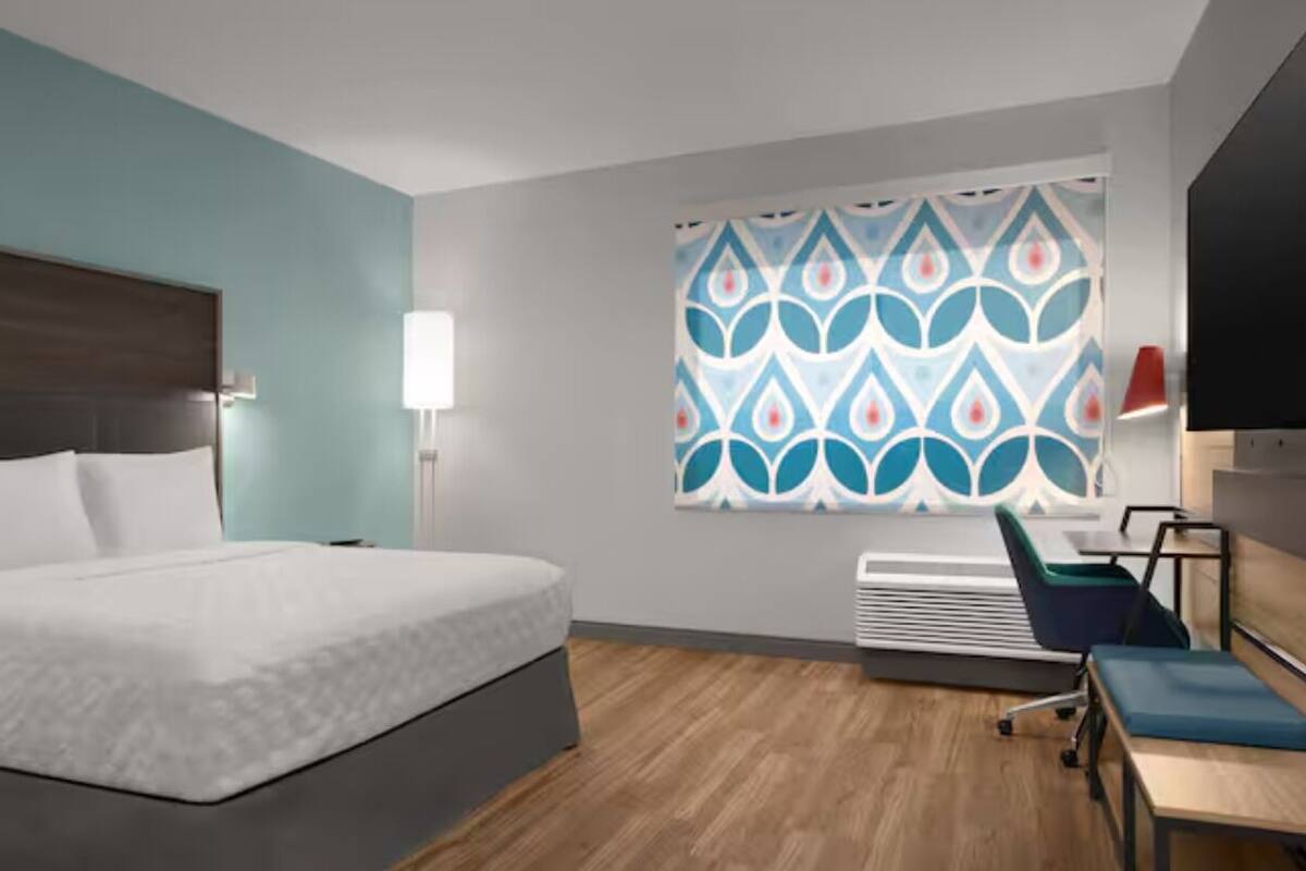 A hotel room with a bed, desk, chair, and a window with patterned curtains.