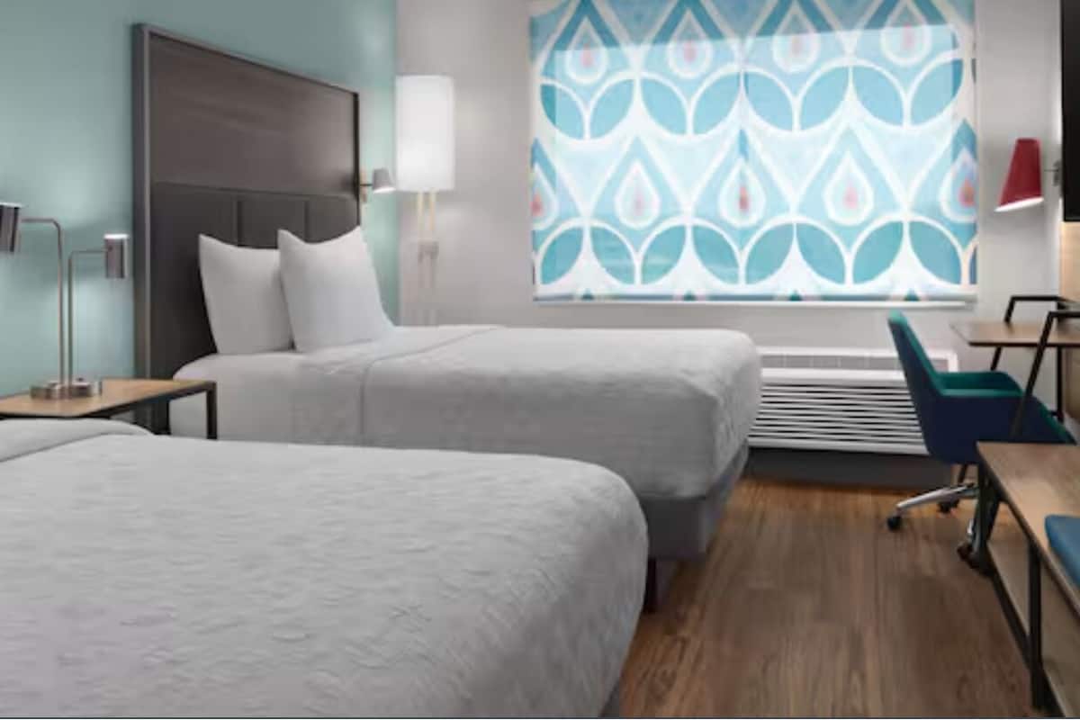 A hotel room with two beds, a desk, a chair, a TV, and a window with a patterned curtain.