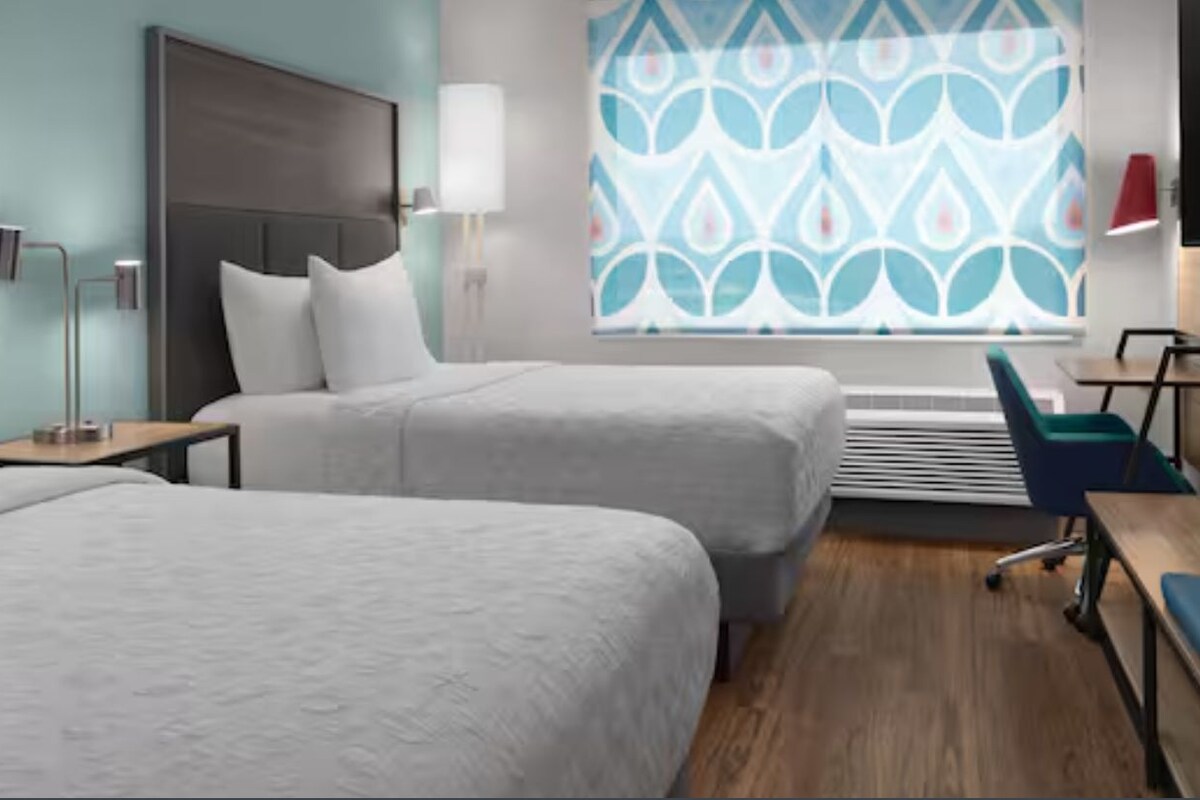Habitación de hotel con dos camas, un escritorio, una silla, un televisor y una ventana con cortina estampada.