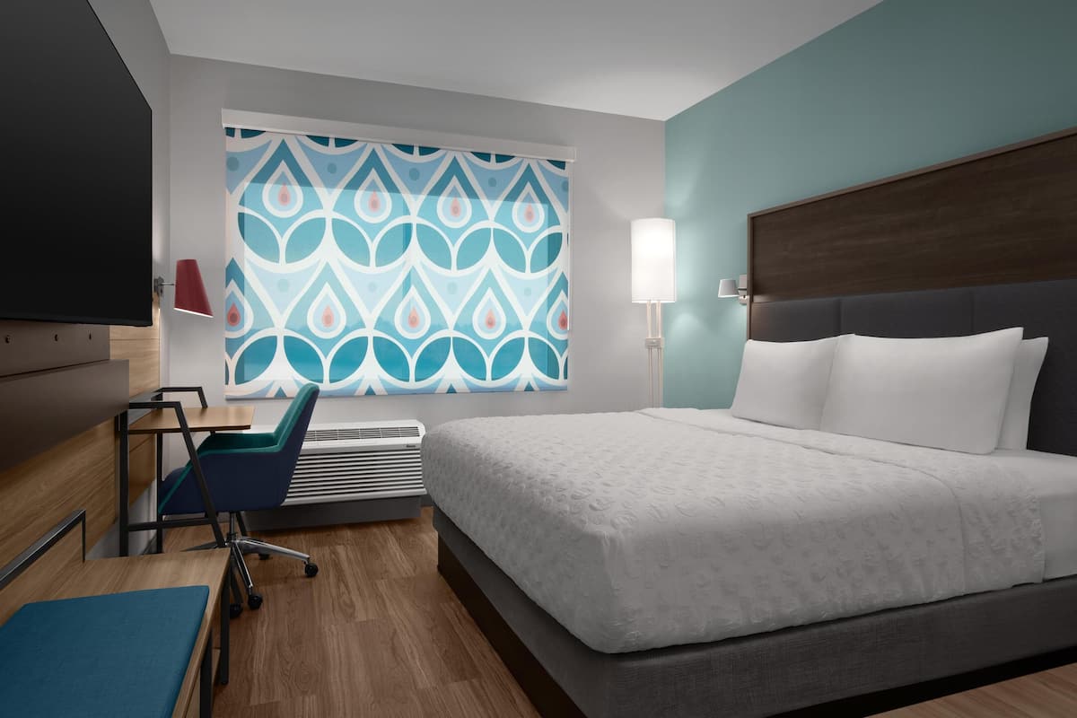 A modern hotel room with a large bed, a desk with a chair, a wall-mounted TV, and a patterned wall.