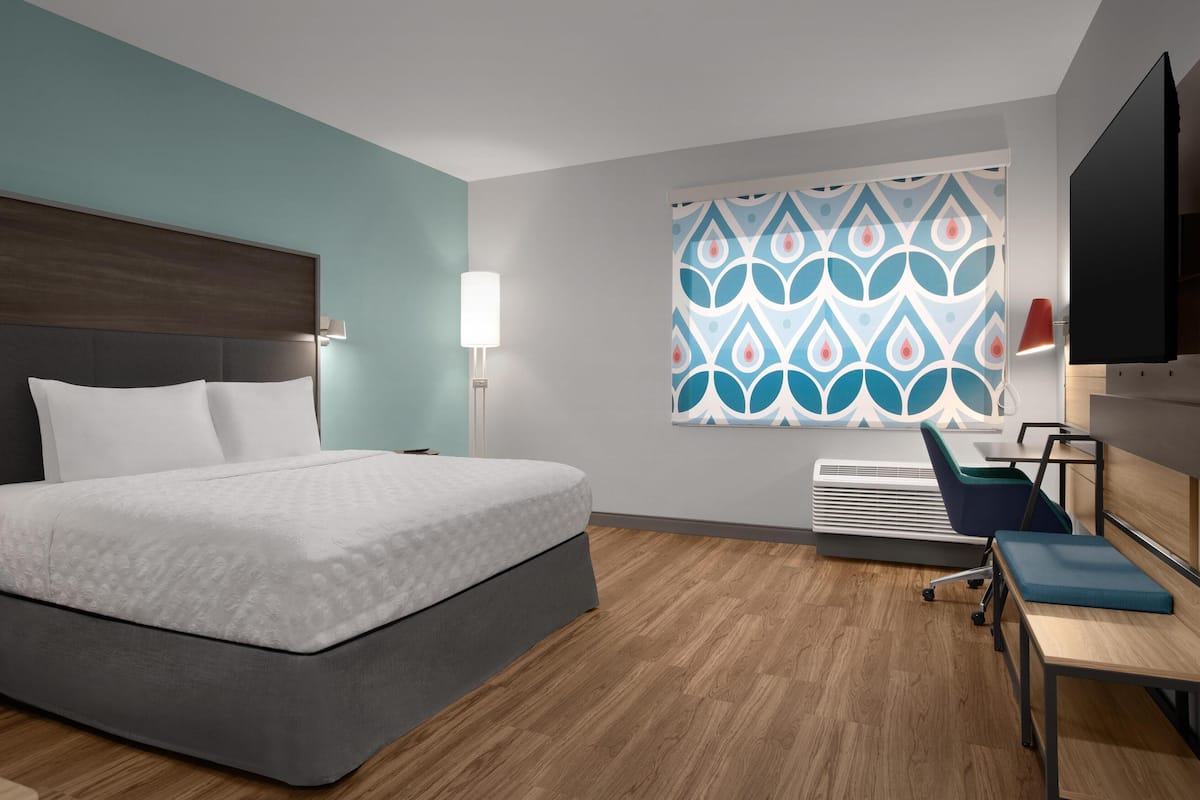 A modern hotel room with a large bed, a desk with a chair, a flat-screen TV, and a patterned window treatment.