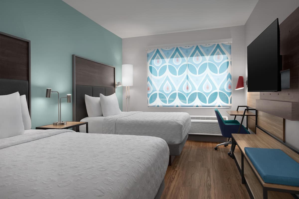 A hotel room with two beds, a desk, a chair, a TV, and a window with a patterned curtain.