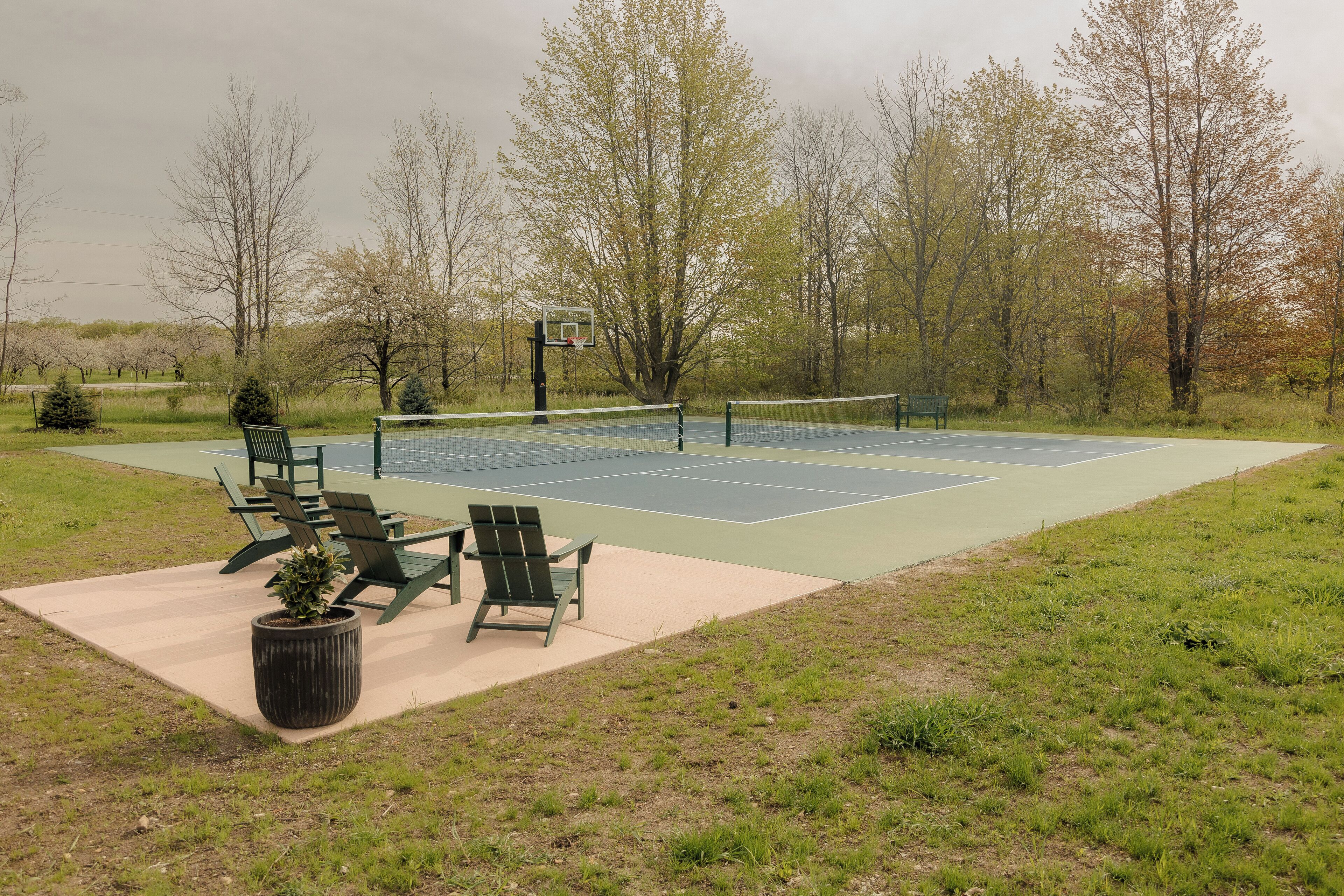 Una cancha de tenis al aire libre con red, rodeada de árboles y una zona de estar verde con sillas y una planta en maceta.