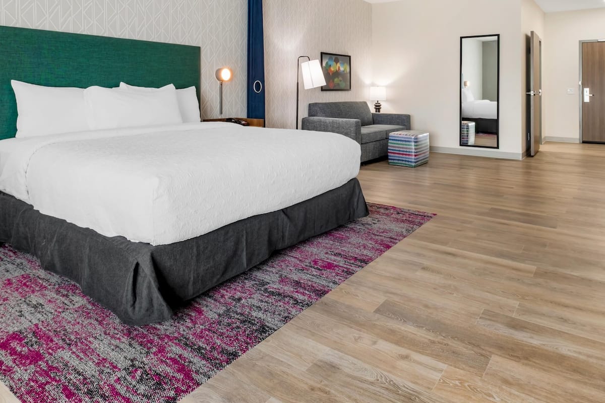 A modern hotel room with a large bed, a desk, and a chair. There is a mirror, a lamp, and a small rug.