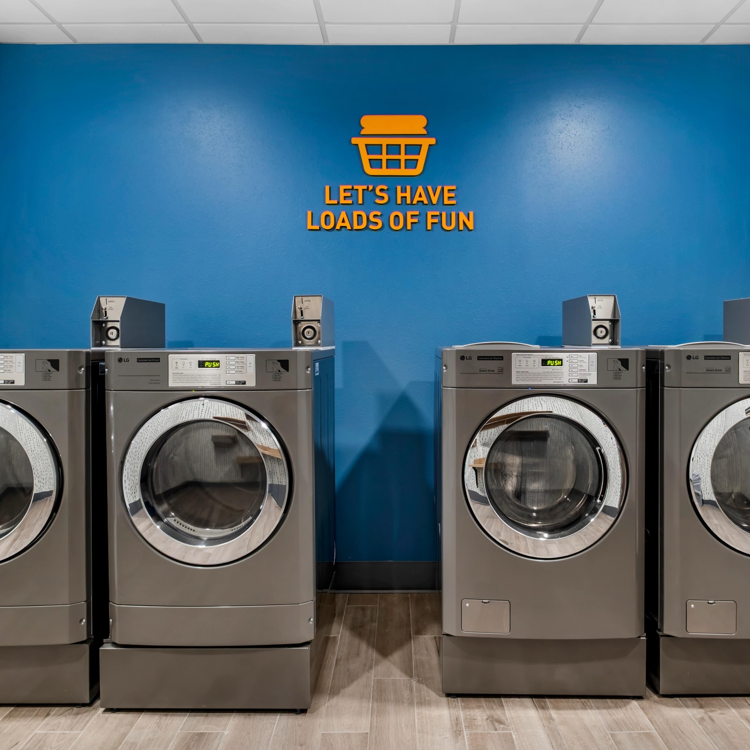 A row of LG front-loading washing machines in a laundry room with a blue wall and a sign that says "LET'S HAVE LOADS OF FUN".
