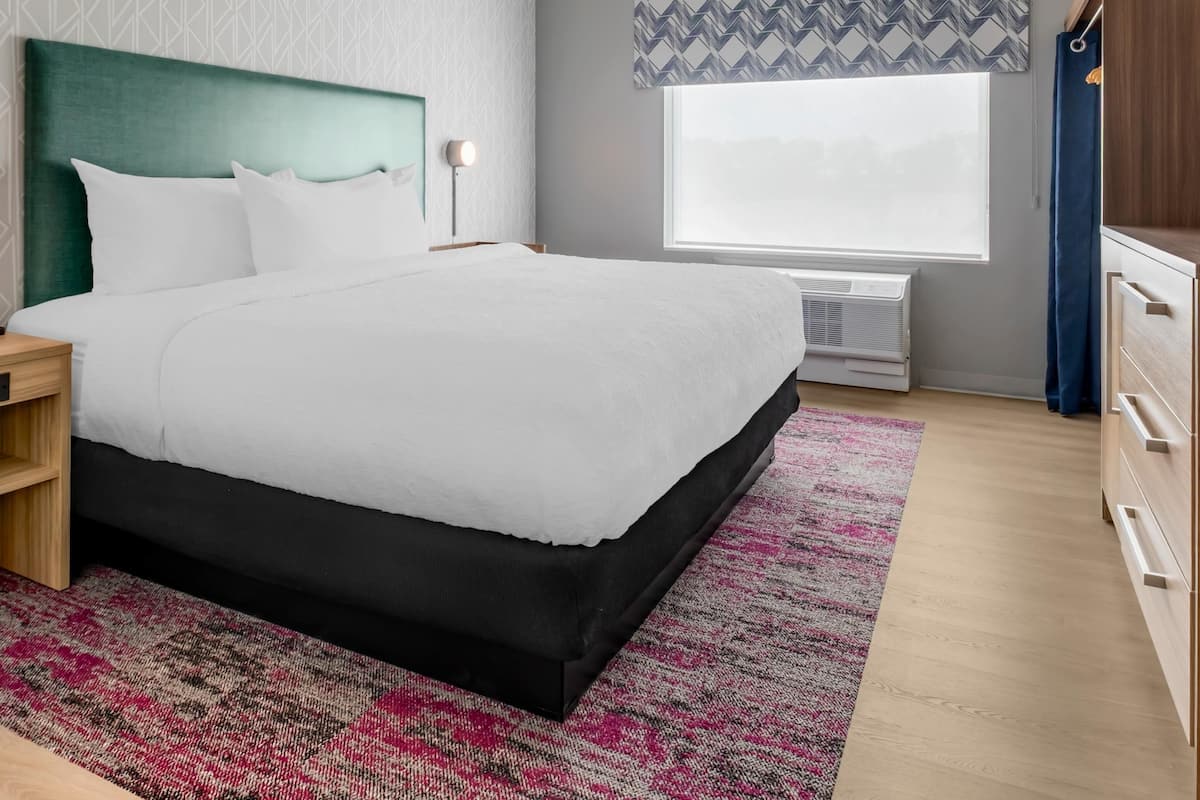 A neatly arranged hotel room with a large bed, a bedside table, a nightstand, a dresser, a window with a view, and a patterned wall.