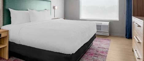 A neatly arranged hotel room with a large bed, a bedside table, a nightstand, a dresser, a window with a view, and a patterned wall.