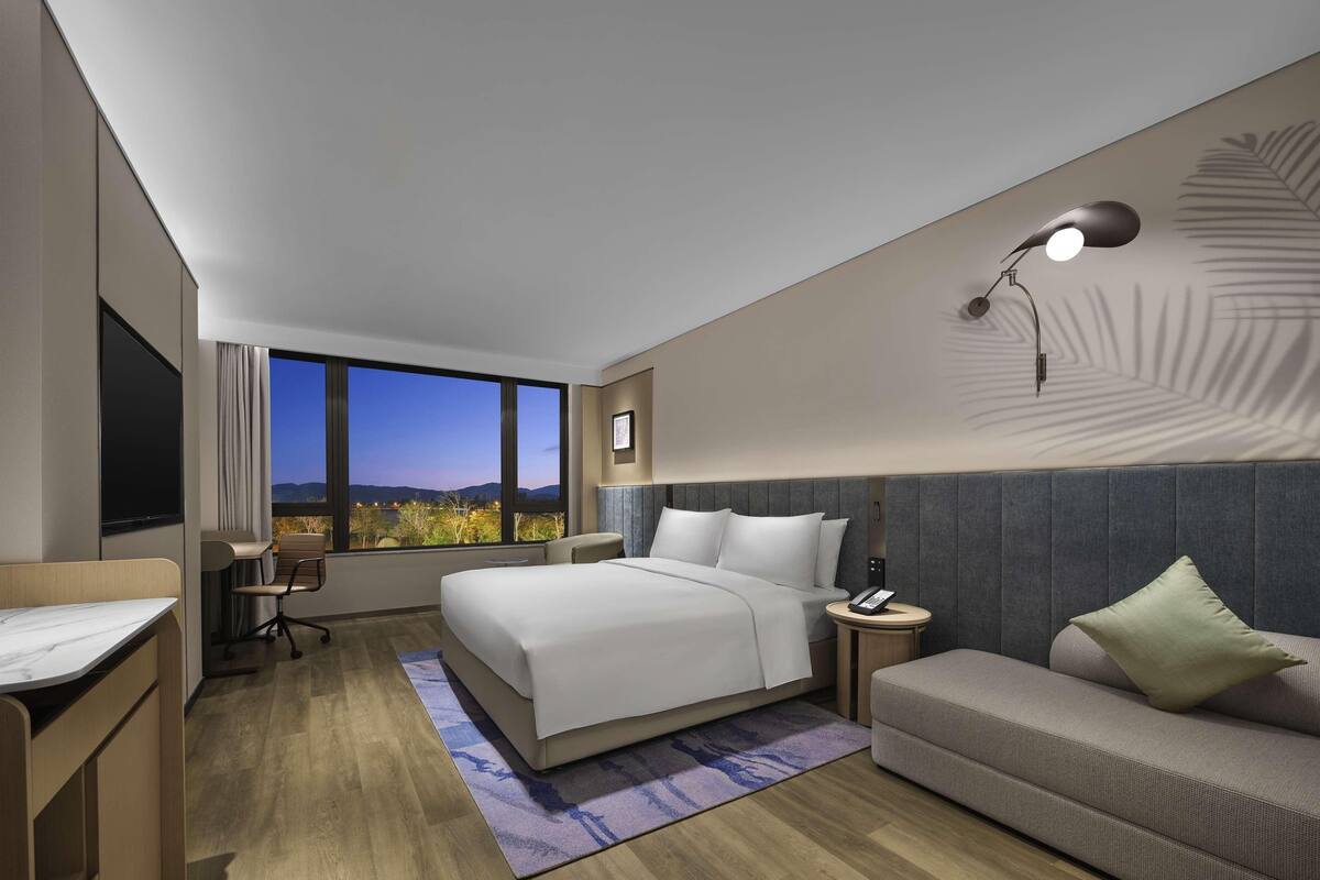 A modern hotel room with a large bed, a sofa, a desk, and a view of the mountains.
