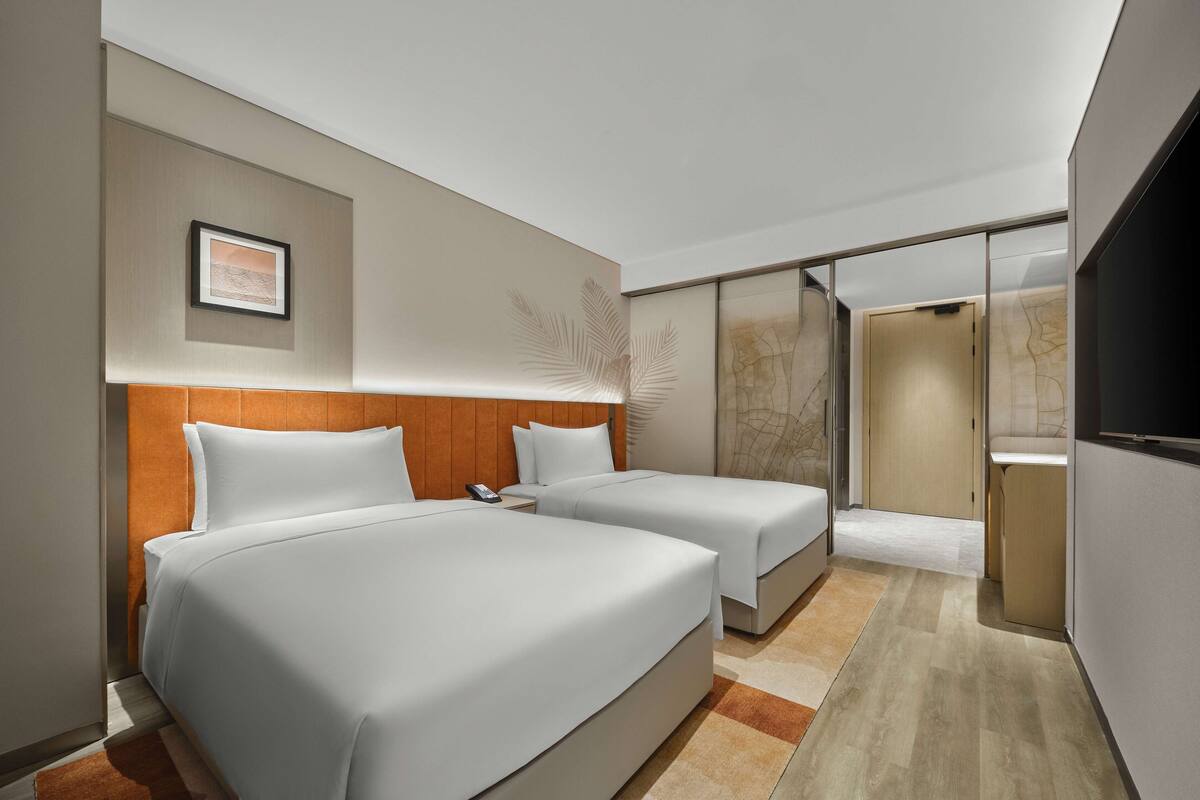 A modern hotel room with two beds, a large window, and a sliding door leading to a bathroom.