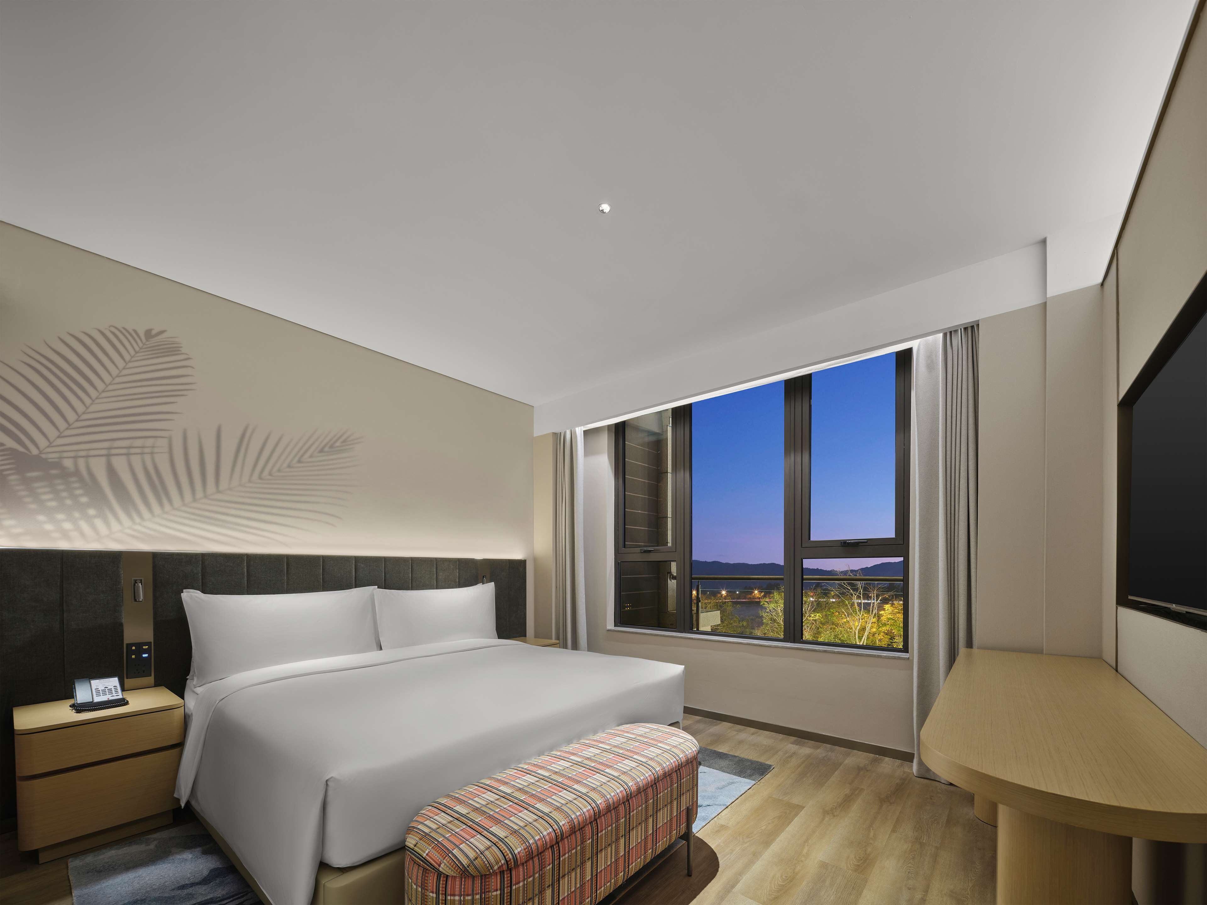 Een moderne hotelkamer met een groot bed, een nachtkastje met telefoon, een bankje en uitzicht op een stadsbeeld door grote ramen.