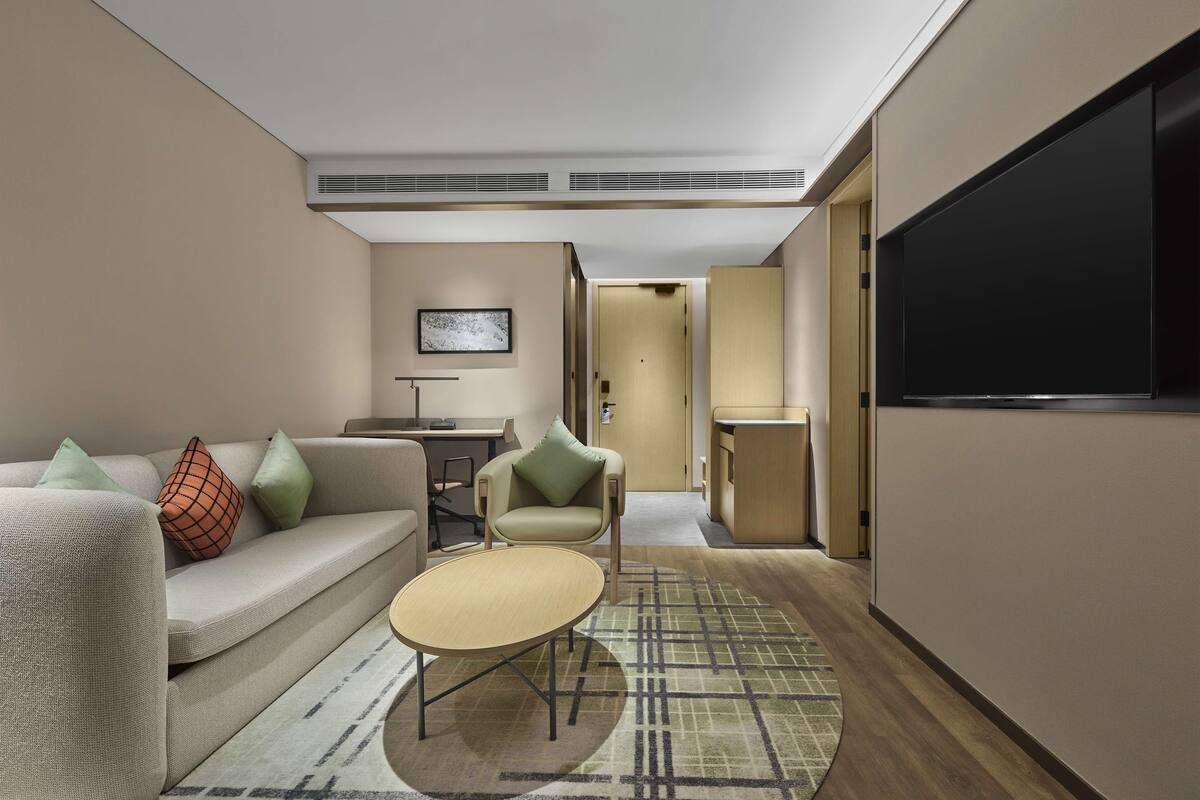 A modern hotel room with a sofa, armchairs, a small table, a TV, and a door leading to another room.