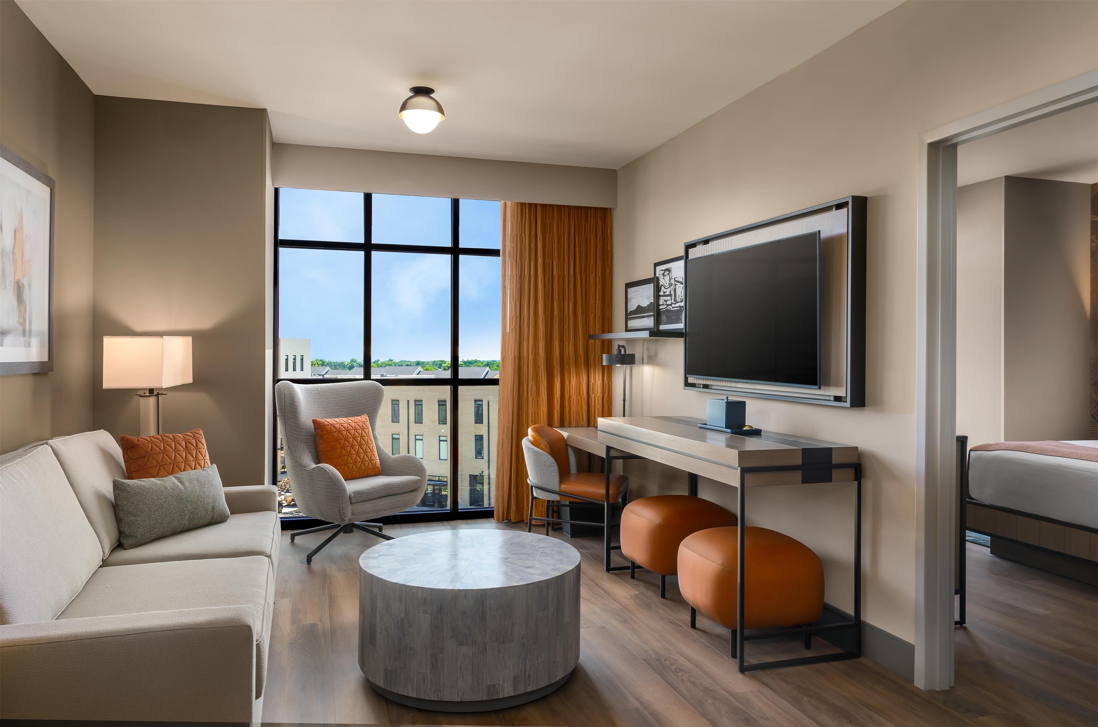 A modern hotel room with a sofa, armchair, round coffee table, desk, and a bed.