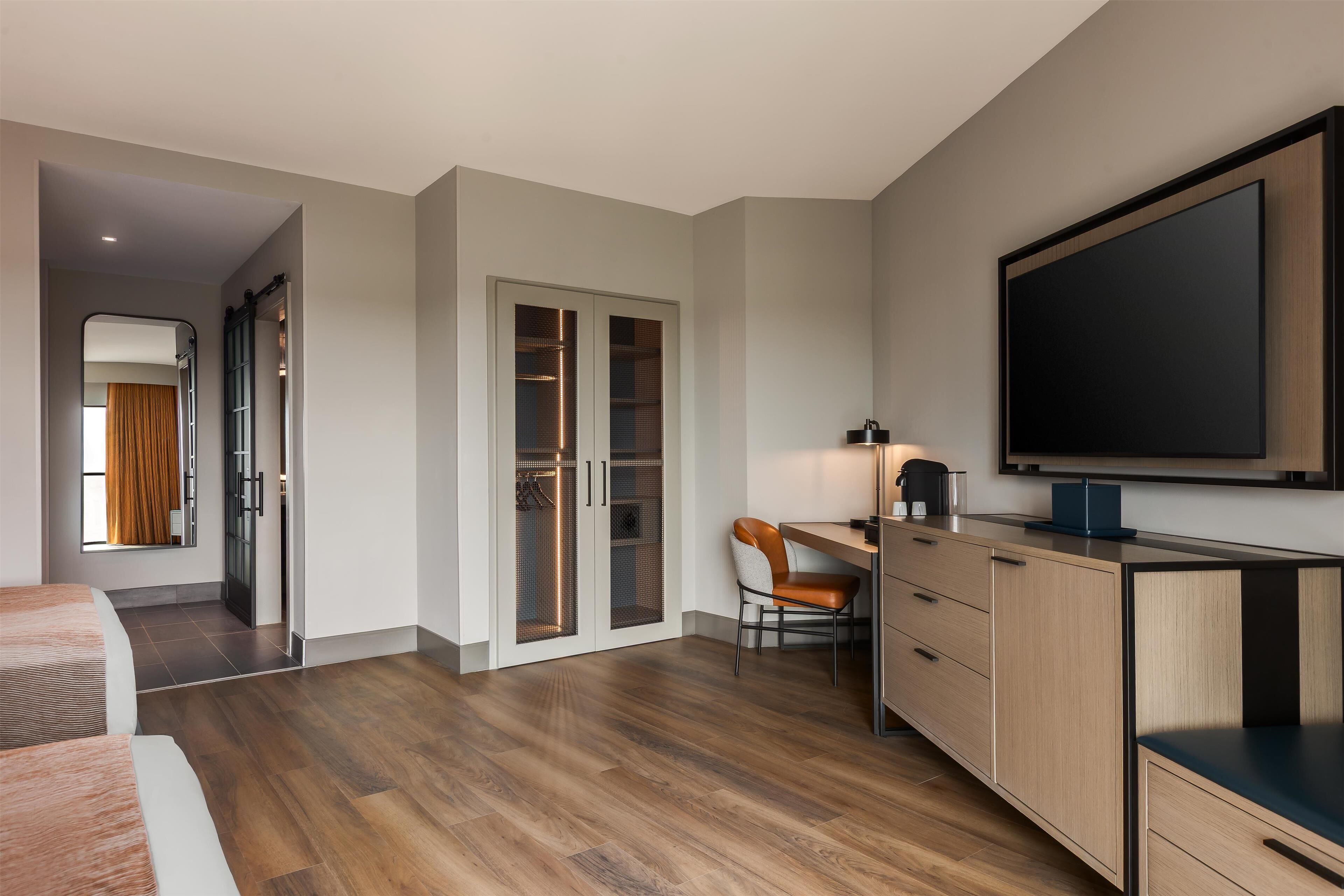 A modern hotel room with a large bed, a desk with a chair, a wardrobe, and a TV mounted on the wall.