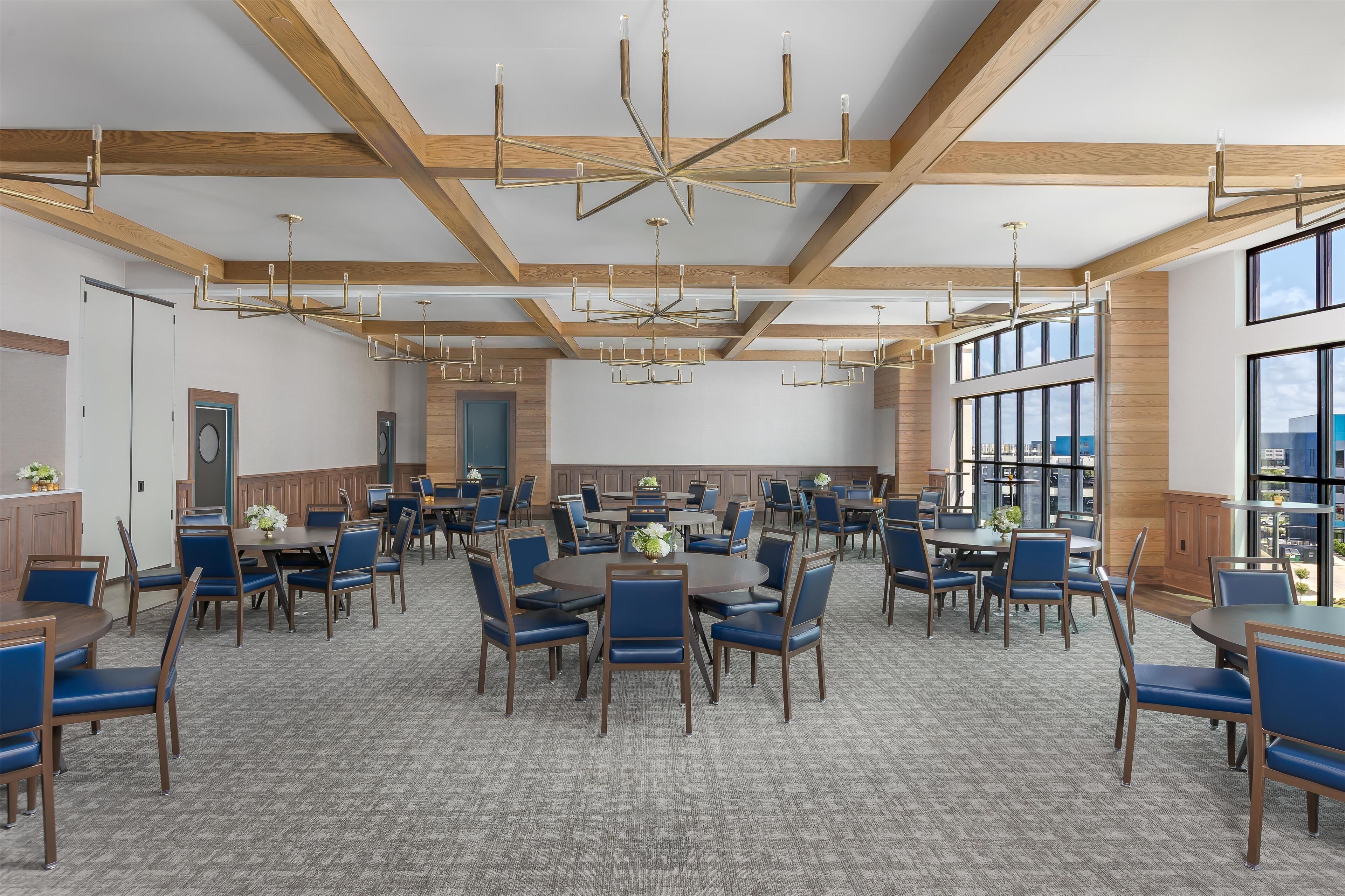 A spacious dining area with multiple tables and chairs, wooden beams on the ceiling, and large windows providing natural light.