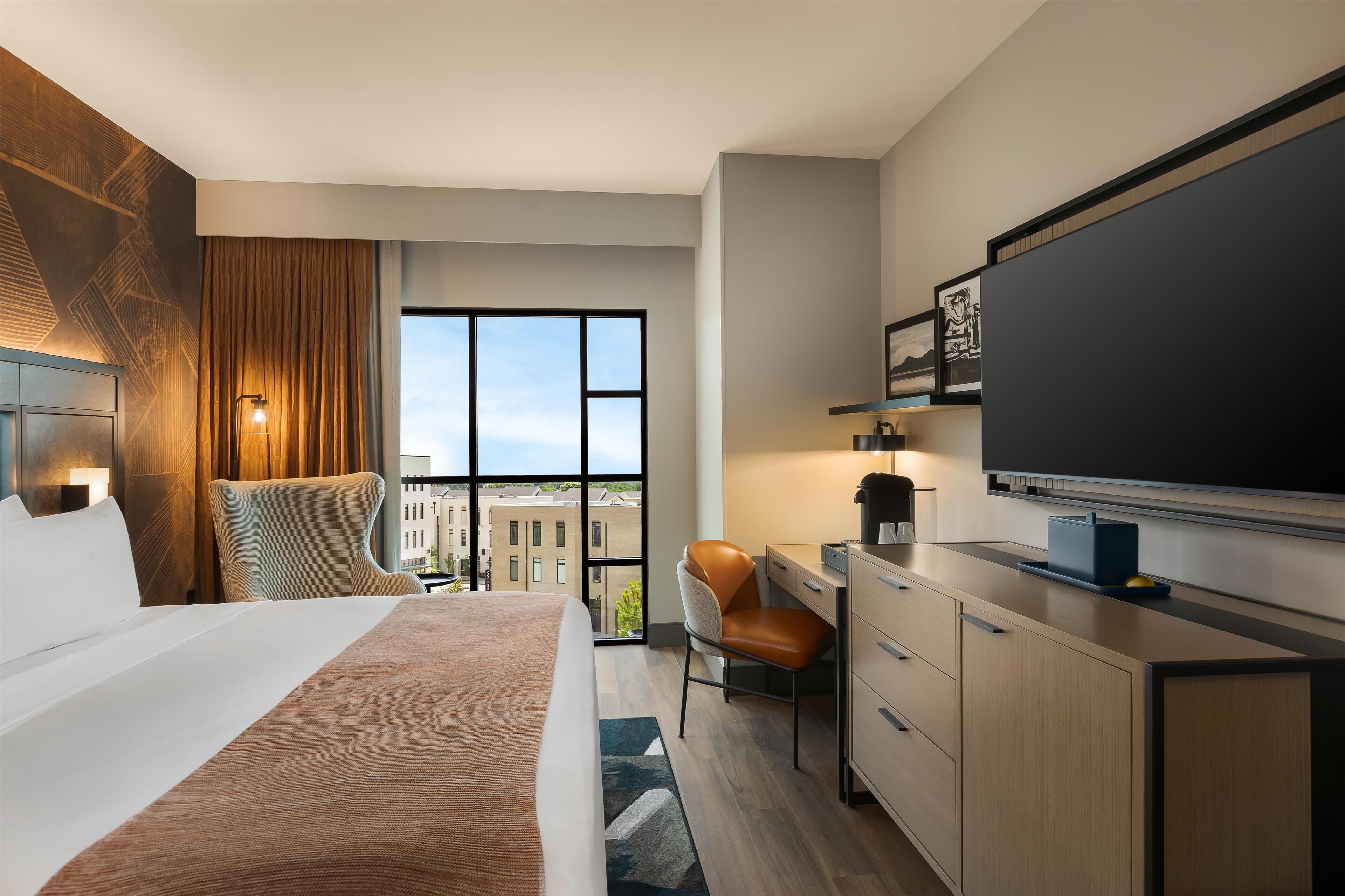 Ett modernt hotellrum med en stor säng, ett skrivbord, en stol, en tv och utsikt över staden genom stora fönster.