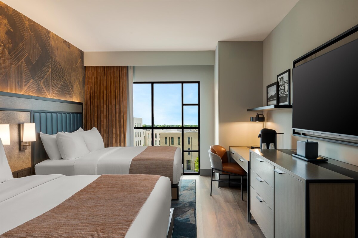 Habitación de hotel con dos camas, un escritorio, una silla, un televisor y un ventanal grande con vistas a la ciudad.