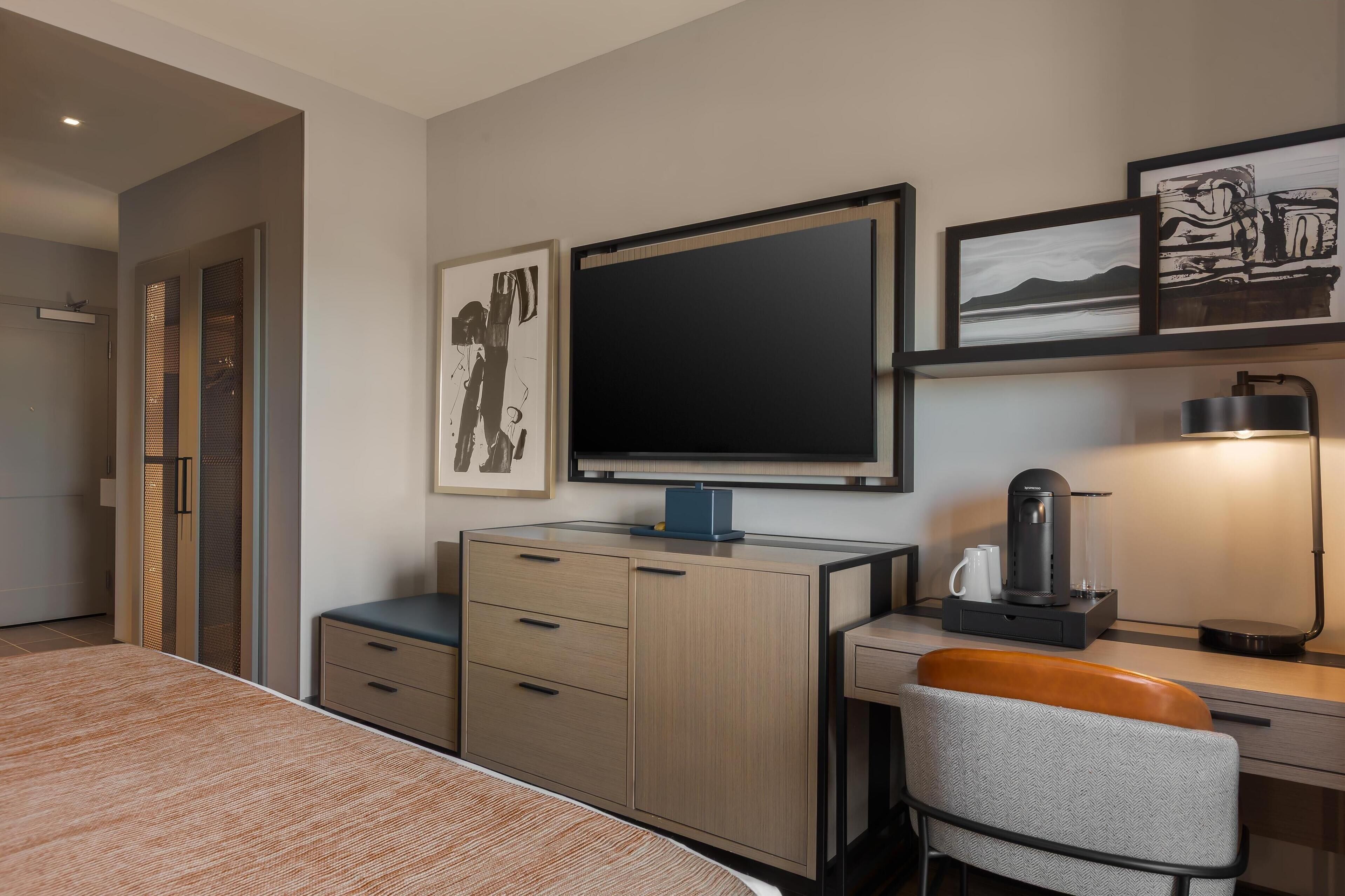 Une chambre d’hôtel moderne équipée d’un lit, d’un bureau avec une machine à café, d’une chaise et d’œuvres d’art accrochées au mur.