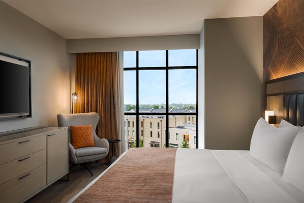 A modern hotel room with a large bed, a TV, a chair, and a view of buildings through large windows. - Hotel Avail Rogers, Tapestry Collection By Hilton (Rogers)