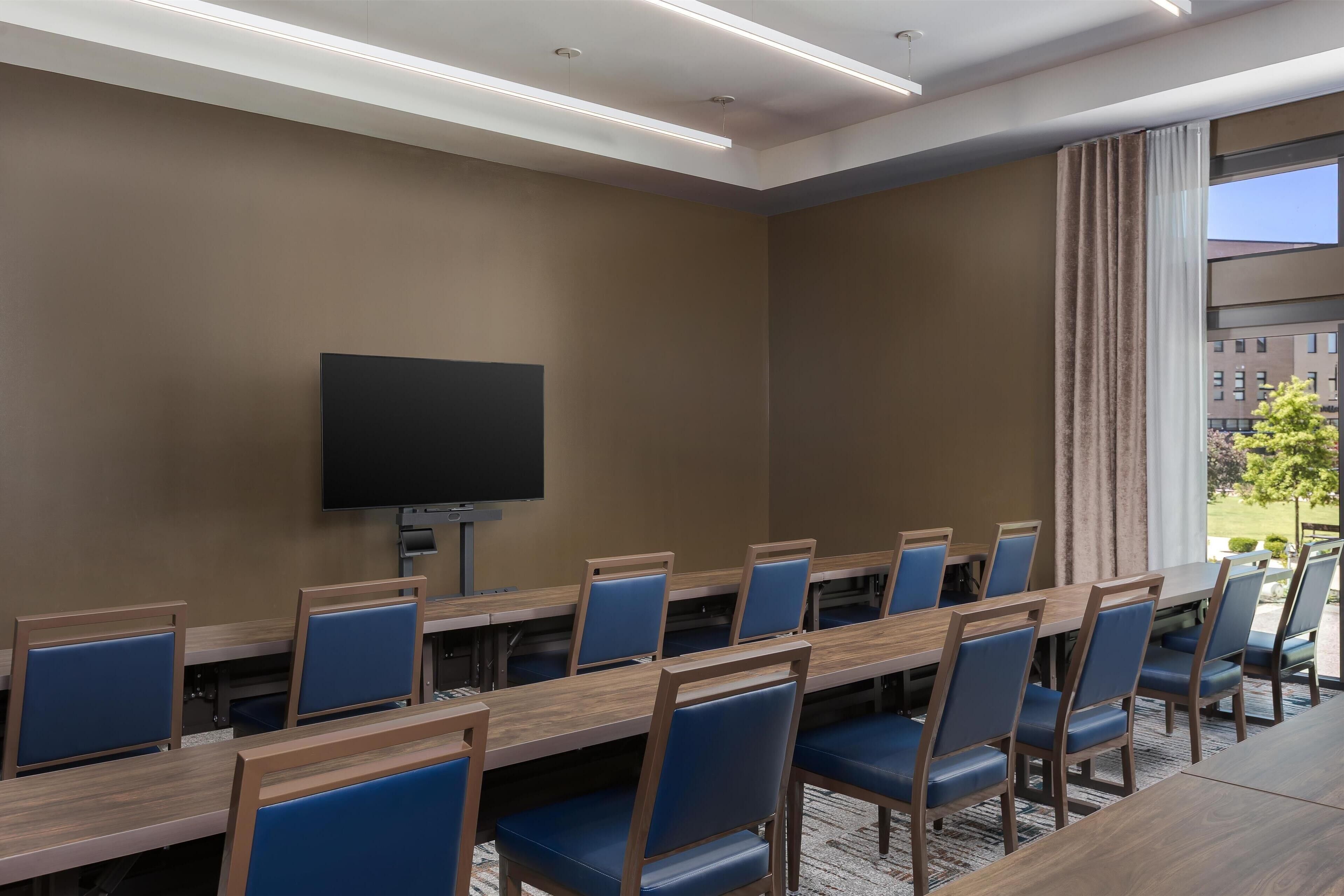 A conference room with a long table, blue chairs, a flat-screen TV, and a large window with curtains.