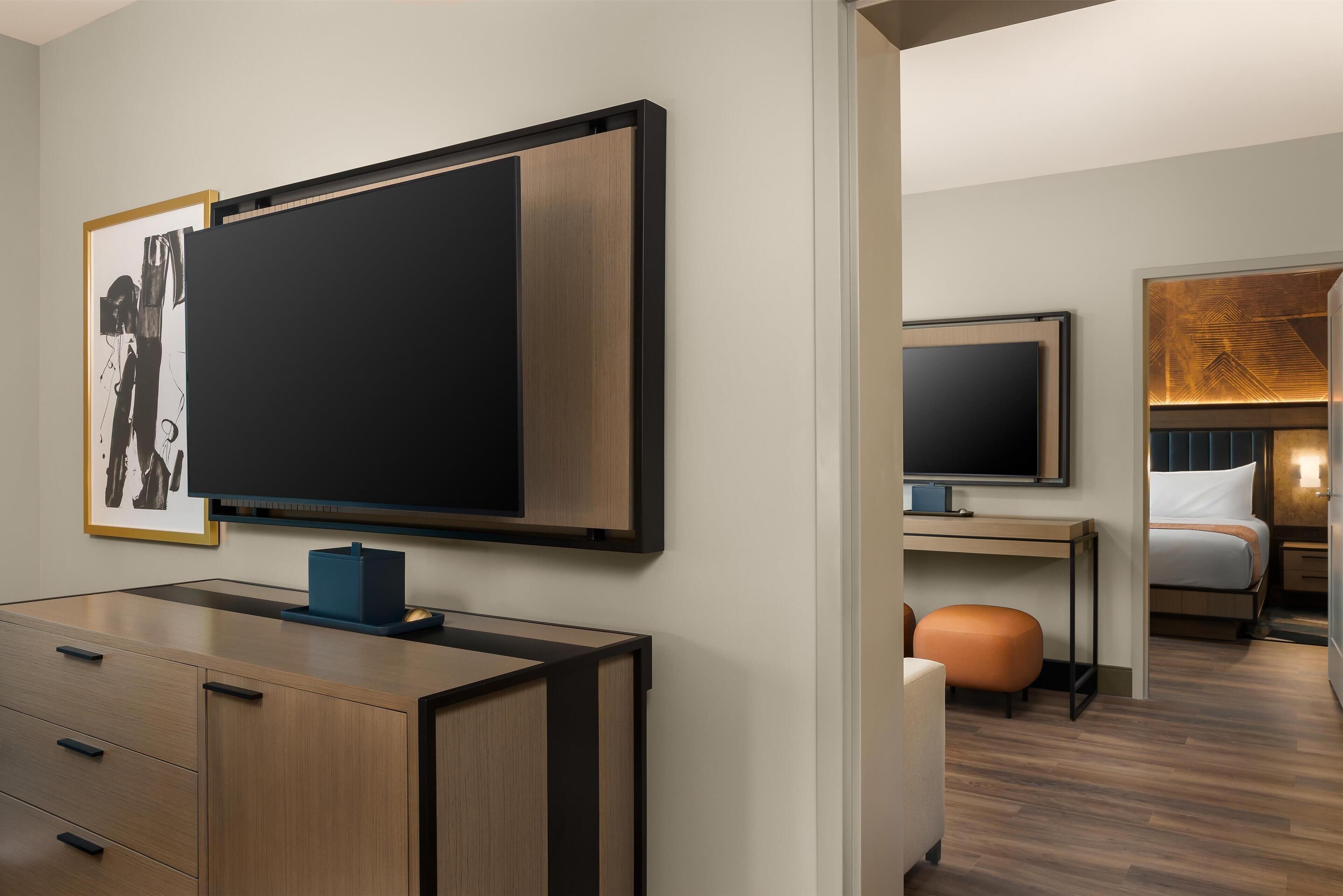 Ein modernes Hotelzimmer mit einem großen Flachbildfernseher an der Wand, einem Holztisch mit Schubladen, einer gemütlichen Sitzecke mit einem orangefarbenen Pouf und einem ordentlich bezogenen Bett mit einem hölzernen Kopfteil.