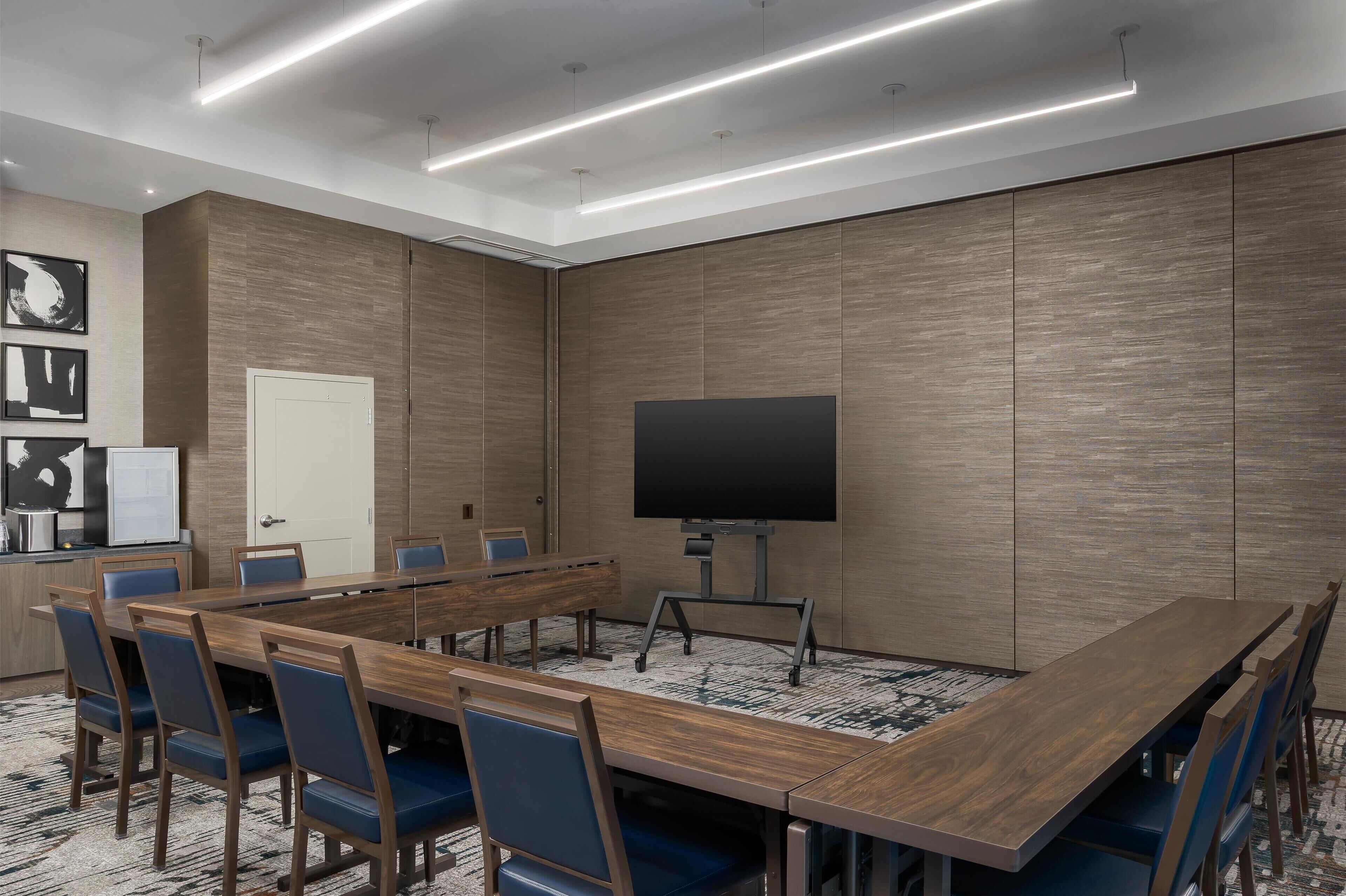 A conference room with a long wooden table, blue chairs, a flat-screen TV, and a patterned carpet.