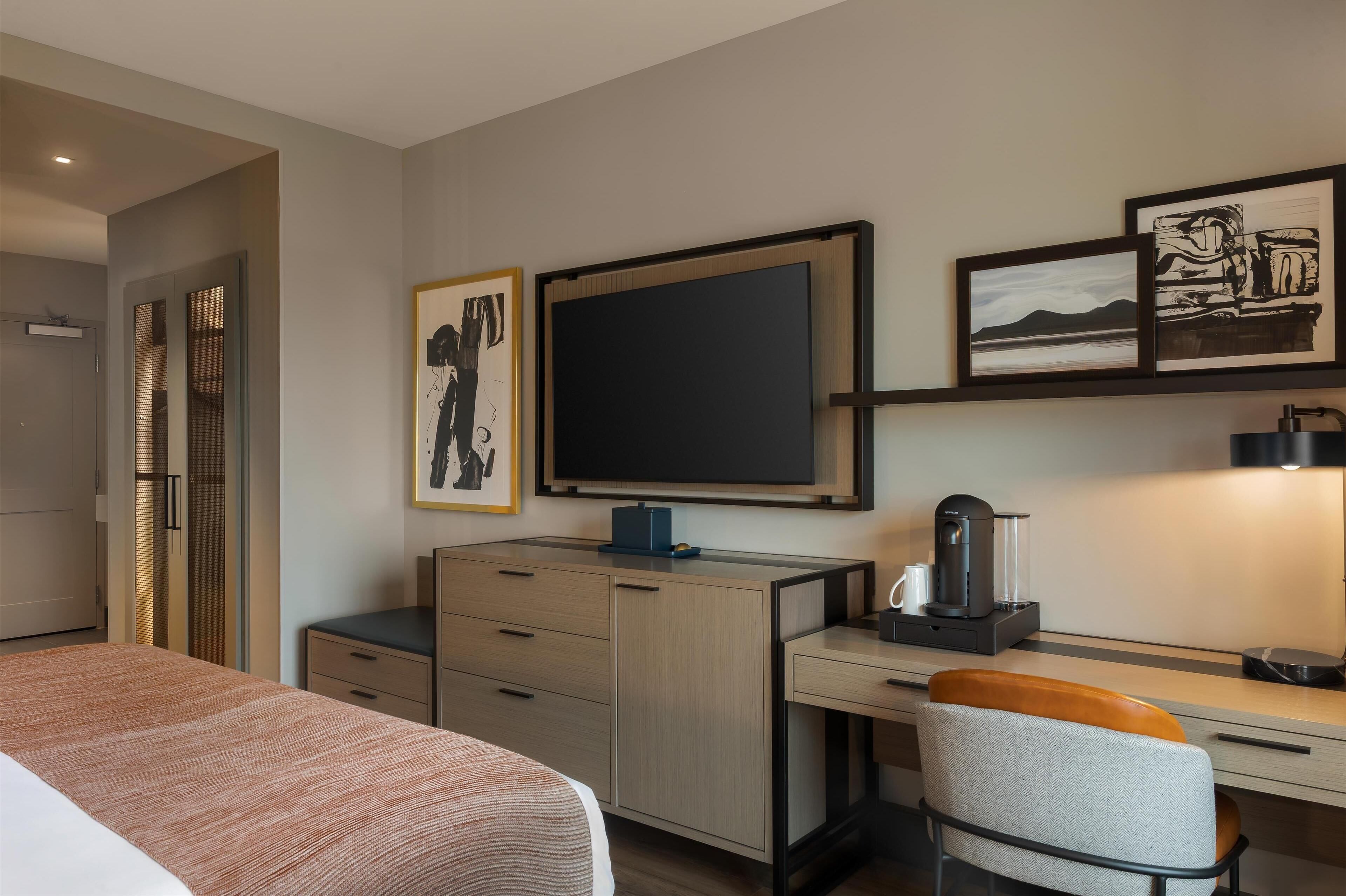 A modern hotel room with a bed, a desk with a computer, a chair, and artwork on the wall.
