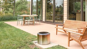A modern outdoor patio with a wooden table and chairs, a metal fire pit, and a glass-enclosed building with sliding doors.