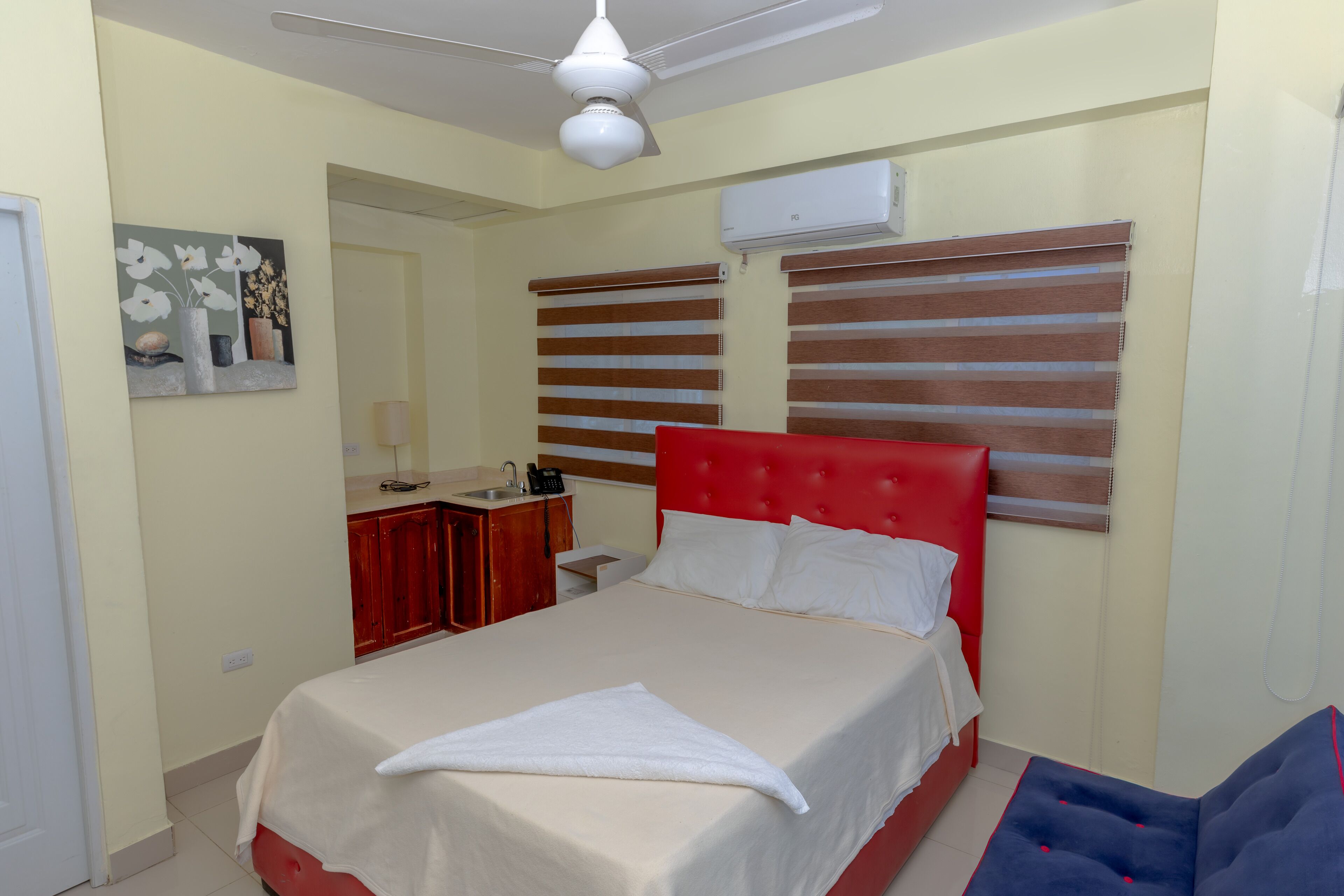 A hotel room with a bed, a red headboard, a bedside table, a mirror, and a window with blinds.