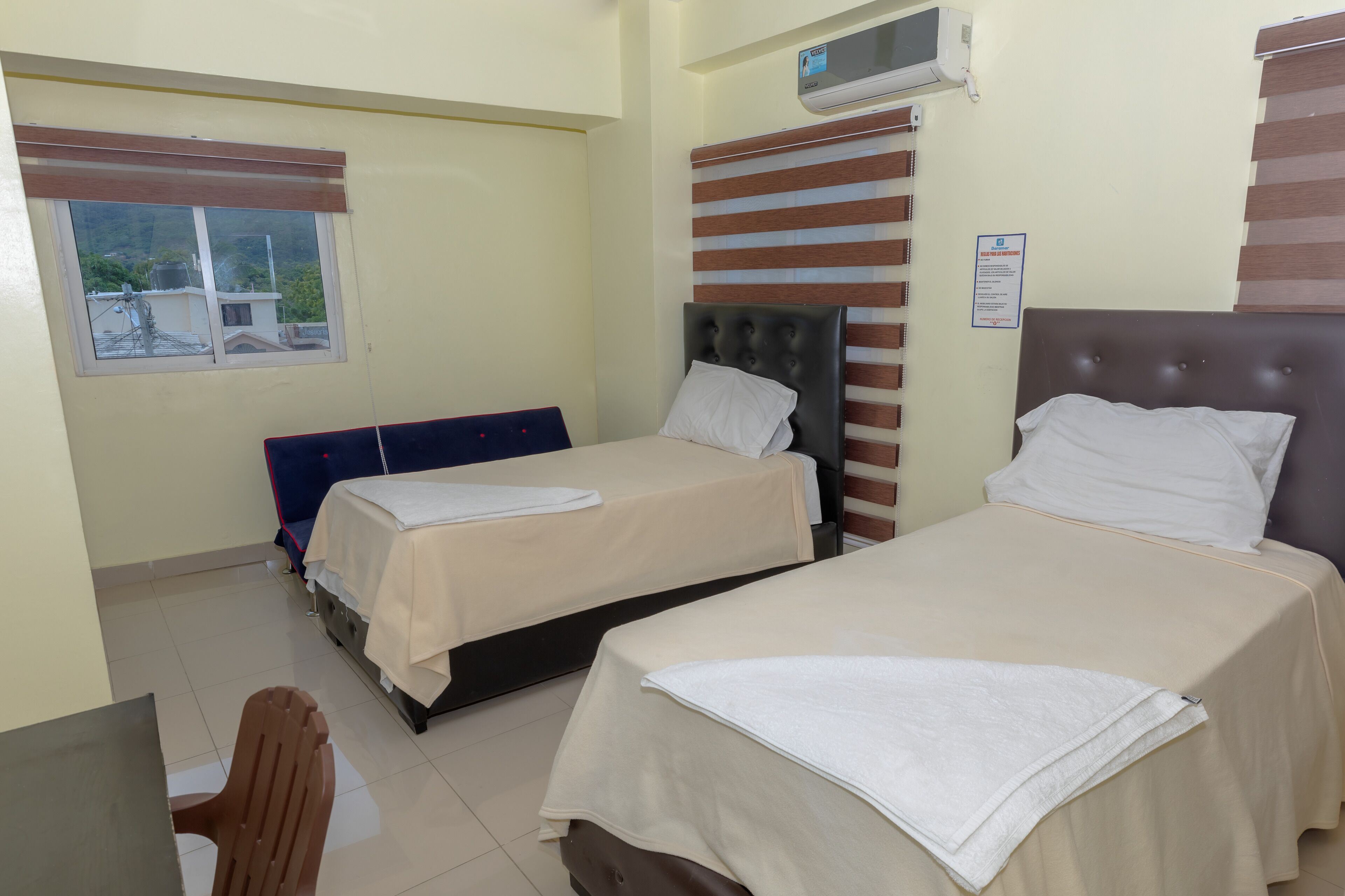 Habitación de hotel con dos camas, una ventana con persianas, un aire acondicionado montado en la pared y una pequeña mesa con una silla.