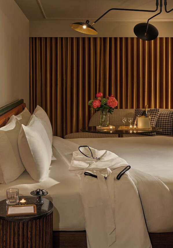 Una habitación de hotel con una cama grande, una mesita de noche con una copa, una vela y una tarjeta enmarcada. Hay una mesa con un jarrón de flores, una botella de champán y copas. La habitación tiene una cortina y una lámpara en el techo.