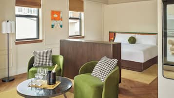 A hotel room with a bed, two green armchairs, a round table with wine glasses, and a large window.