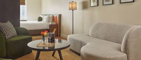 A hotel room with a green armchair, a grey sofa, a round coffee table with a vase of flowers, a bedside table with a bottle of champagne, and a bed with white linens.