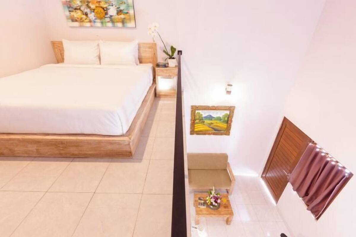A bedroom with a bed, a nightstand, a small table with a vase, a sofa, and a painting on the wall.