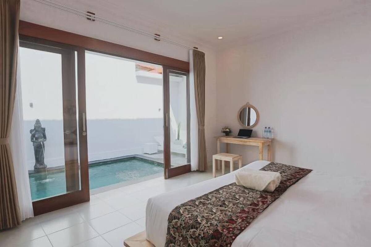 A bedroom with a bed, a desk, a chair, a mirror, and a view of a pool.
