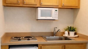 A compact kitchen with wooden cabinets, a white microwave, a sink, and a small refrigerator. - Queens Lodge Motel (West Palm Beach)