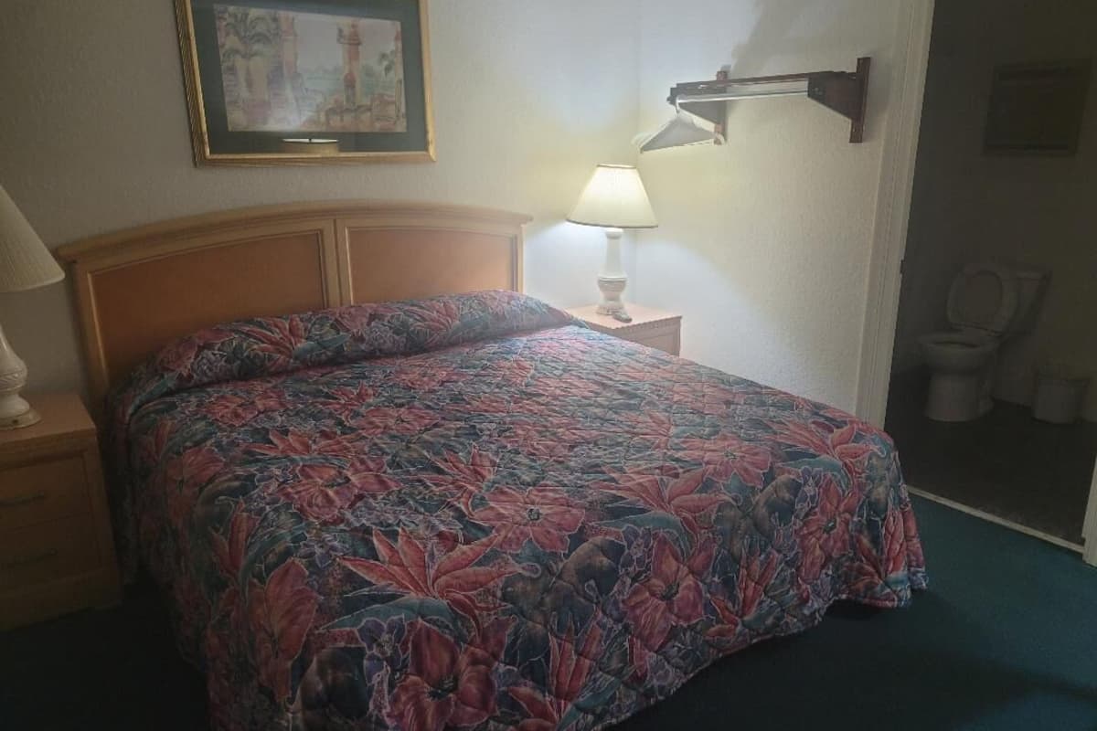 Ein Schlafzimmer mit blumigem Bettüberwurf, einer Nachttischlampe, einem gerahmten Bild an der Wand und einer Toilette in der Ecke.
