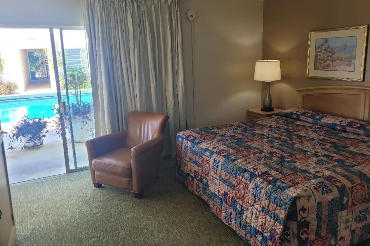 Ein Hotelzimmer mit Bett, Sessel, Lampe und Blick auf einen Swimmingpool.