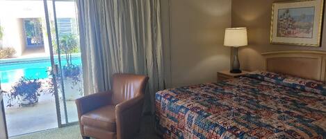 A hotel room with a bed, a chair, a lamp, and a view of a swimming pool.