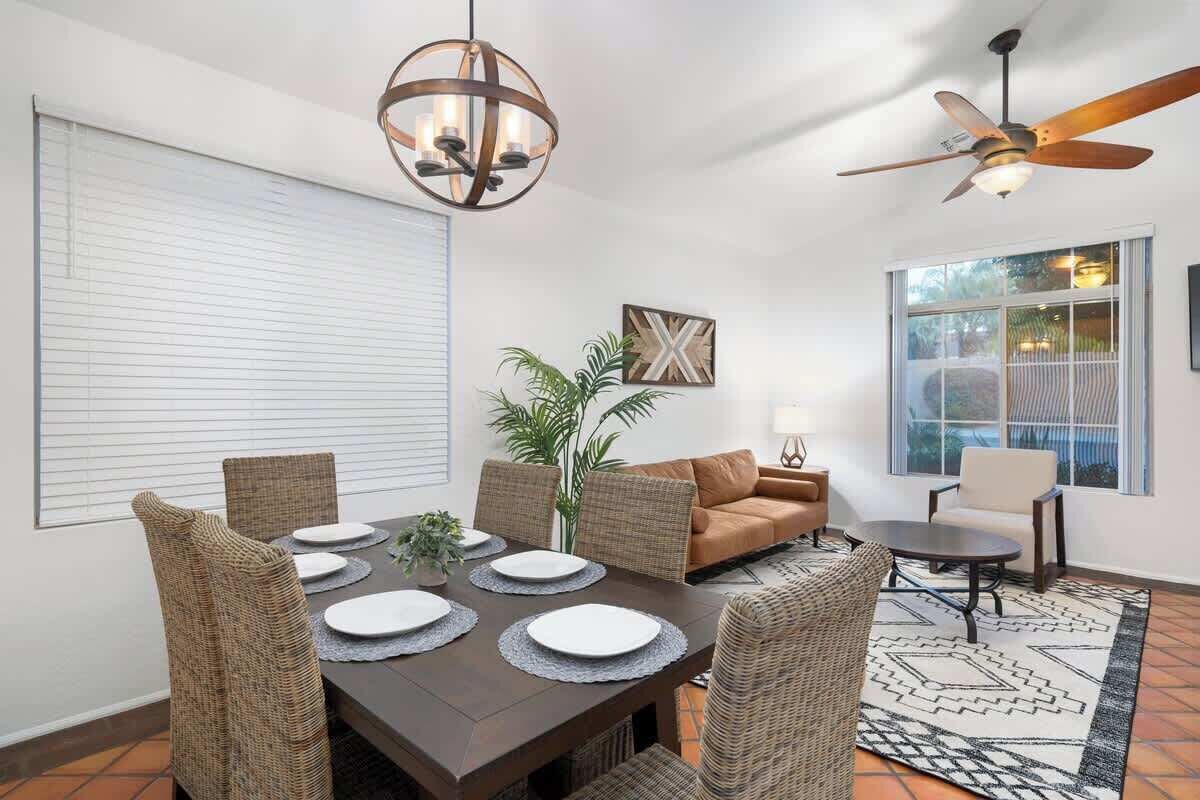 A dining area with a dark wooden table set for four, surrounded by wicker chairs. There is a ceiling fan, a large window with blinds, and a potted plant.