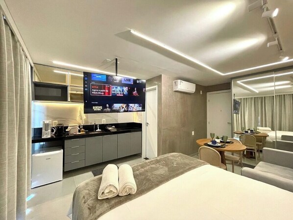 A modern hotel room with a bed, a TV mounted on the wall, a small kitchenette, and a dining area with a table and chairs. - RN STUDIO - APTO 417 (Maceió)